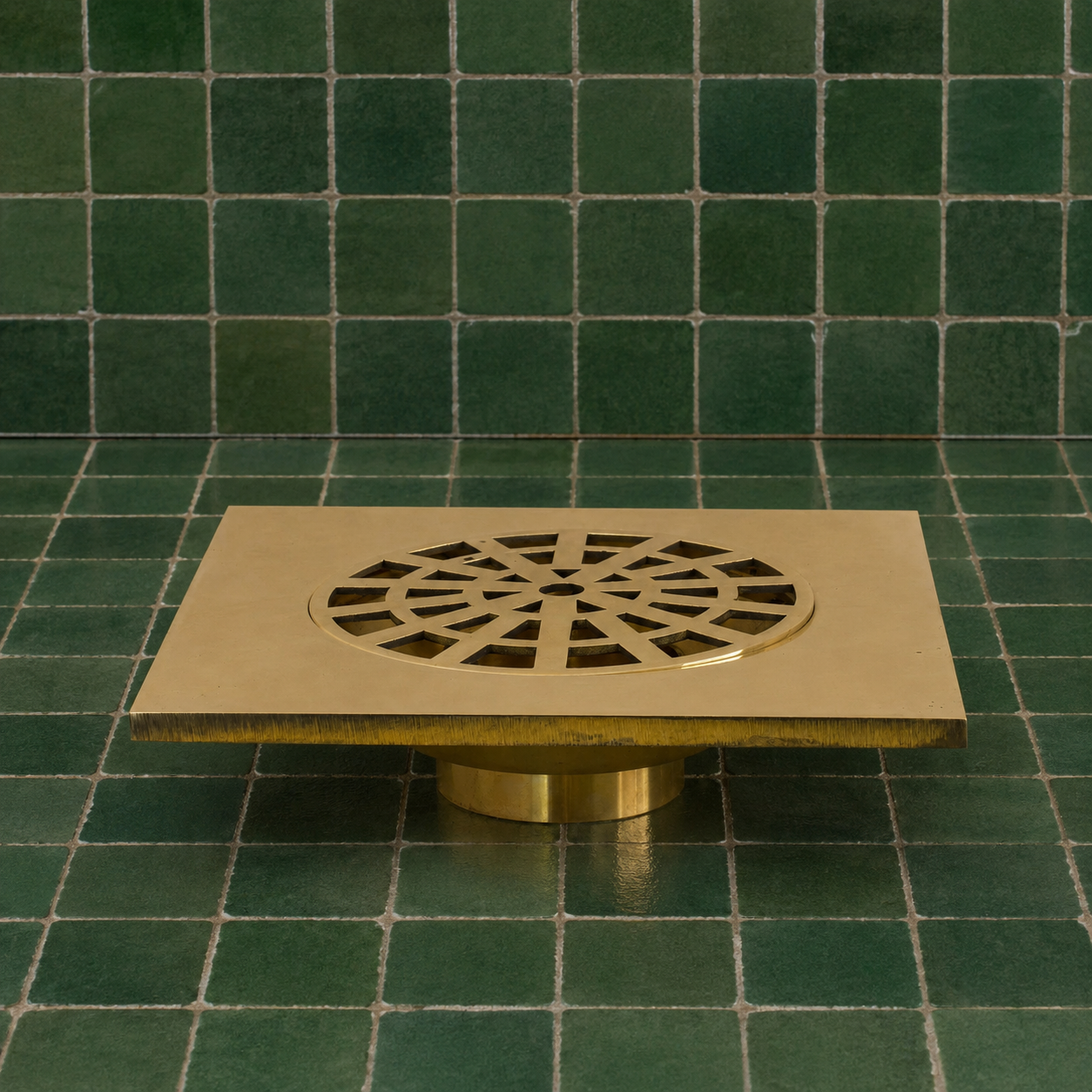A square brass floor drain with a circular cutout and decorative radial pattern, mounted on a cylindrical base against a green tiled wall and floor.