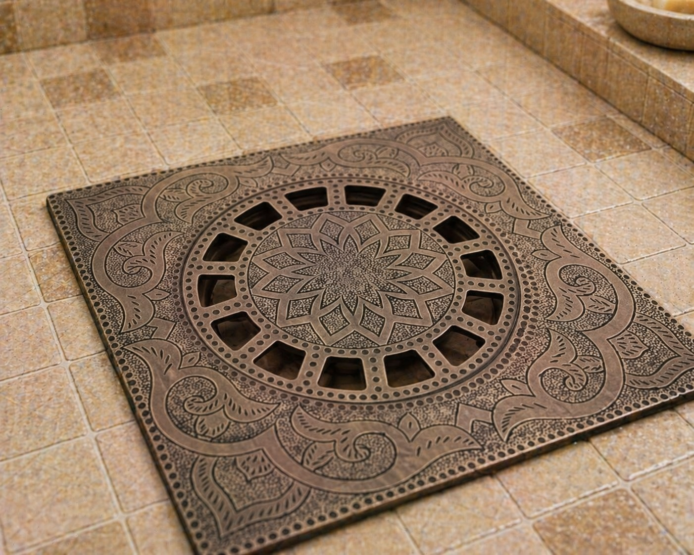 An ornate square metal drain cover with intricate embossed floral and geometric patterns, shown installed on a beige tiled floor.