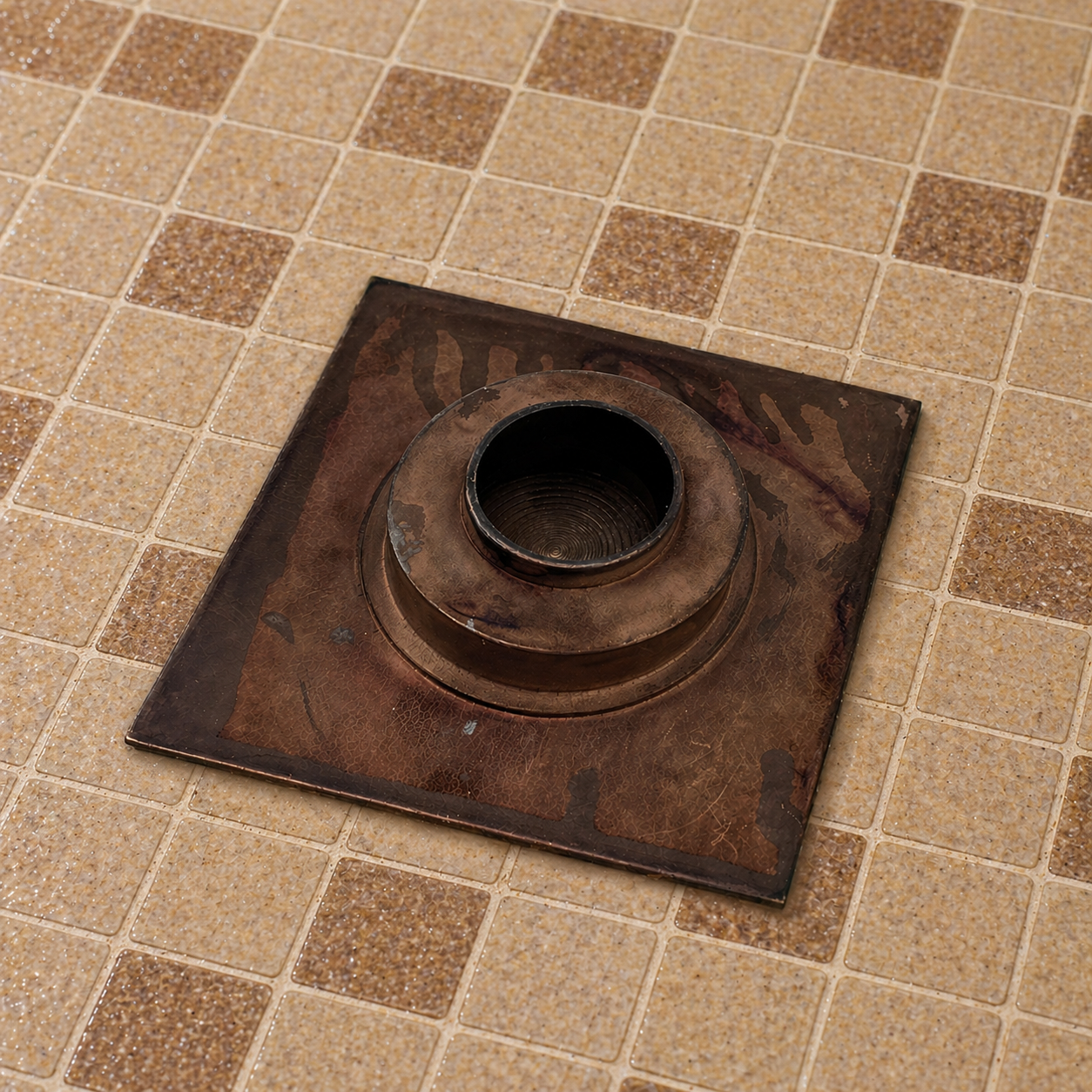 A square metal drain cover with a circular opening, shown placed on a tiled floor.