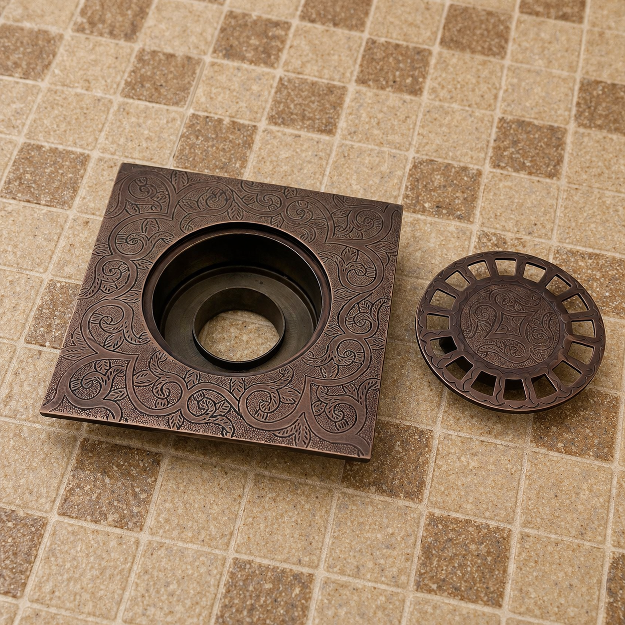 A square bronze-finished drain cover with an embossed floral scroll pattern, shown next to its round drain cap on a beige tiled floor.