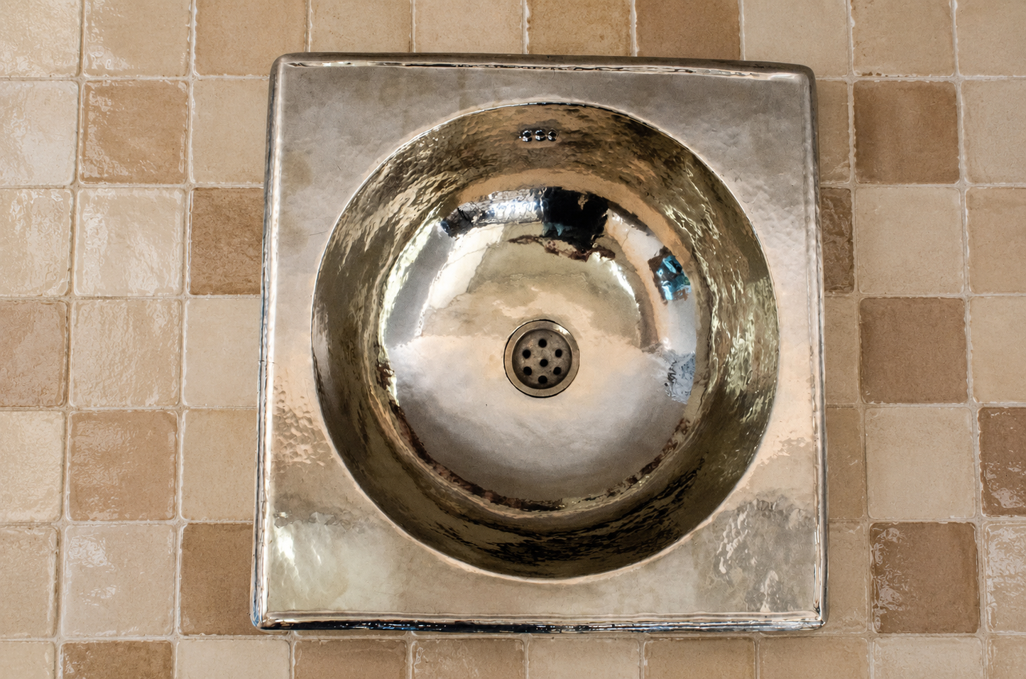 A square metallic sink with a circular basin and central drain, mounted against a beige tiled wall.