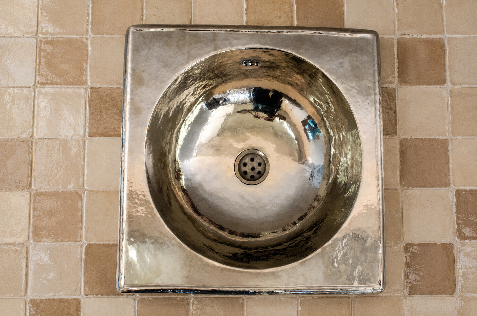 A square metallic sink with a circular basin and central drain, mounted against a beige tiled wall.