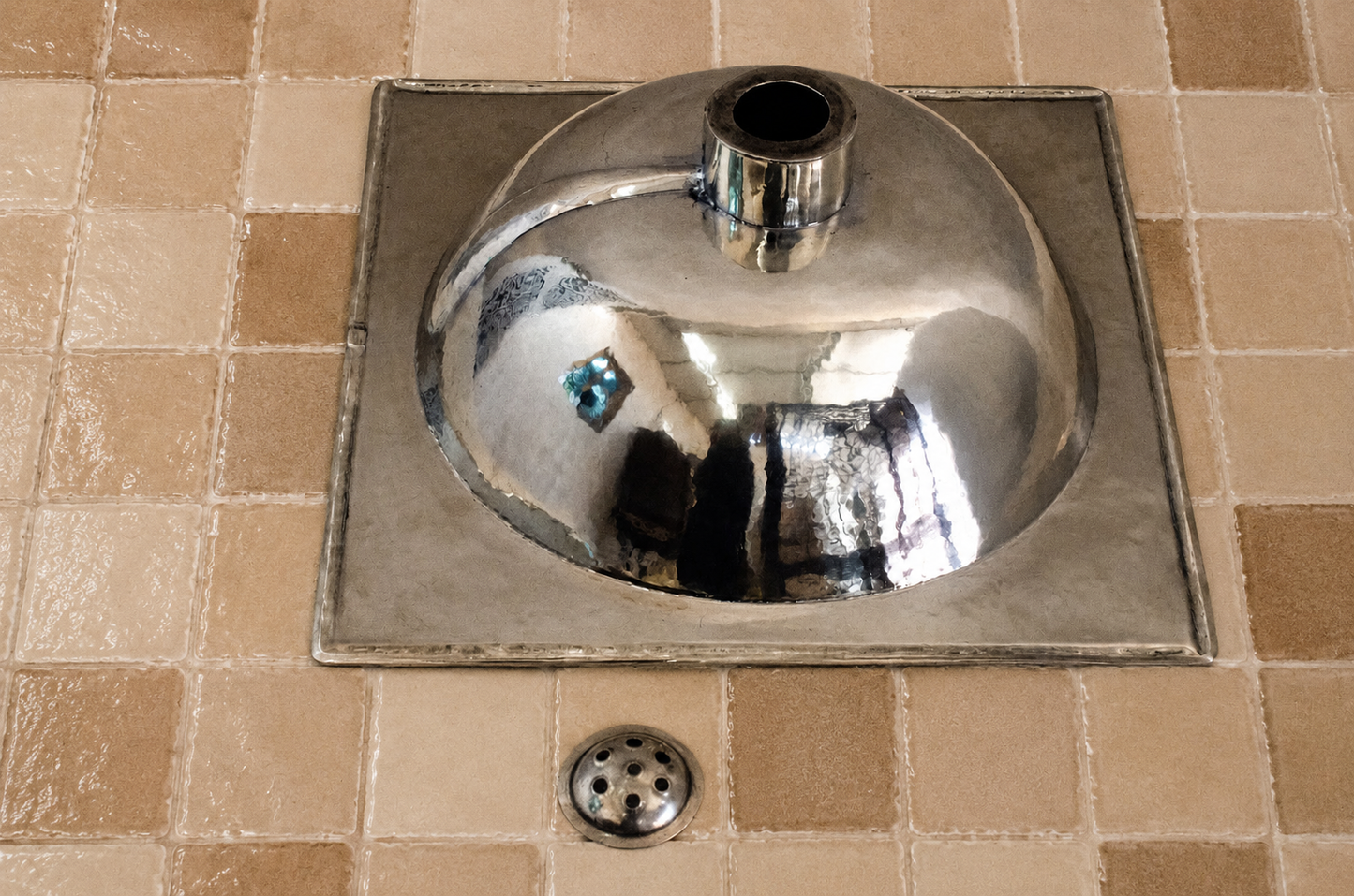 A polished chrome wall-mounted sink basin with a square metal surround and a matching drain, installed on beige tiled wall and floor.