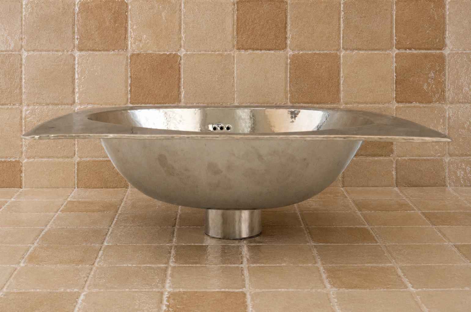A modern stainless steel freestanding bathtub with a wide flared rim, shown against a beige tiled wall and floor.