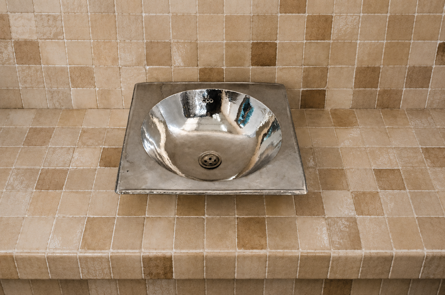 A polished chrome vessel sink basin mounted on a square brushed metal plate, set into a beige tiled countertop and backsplash.