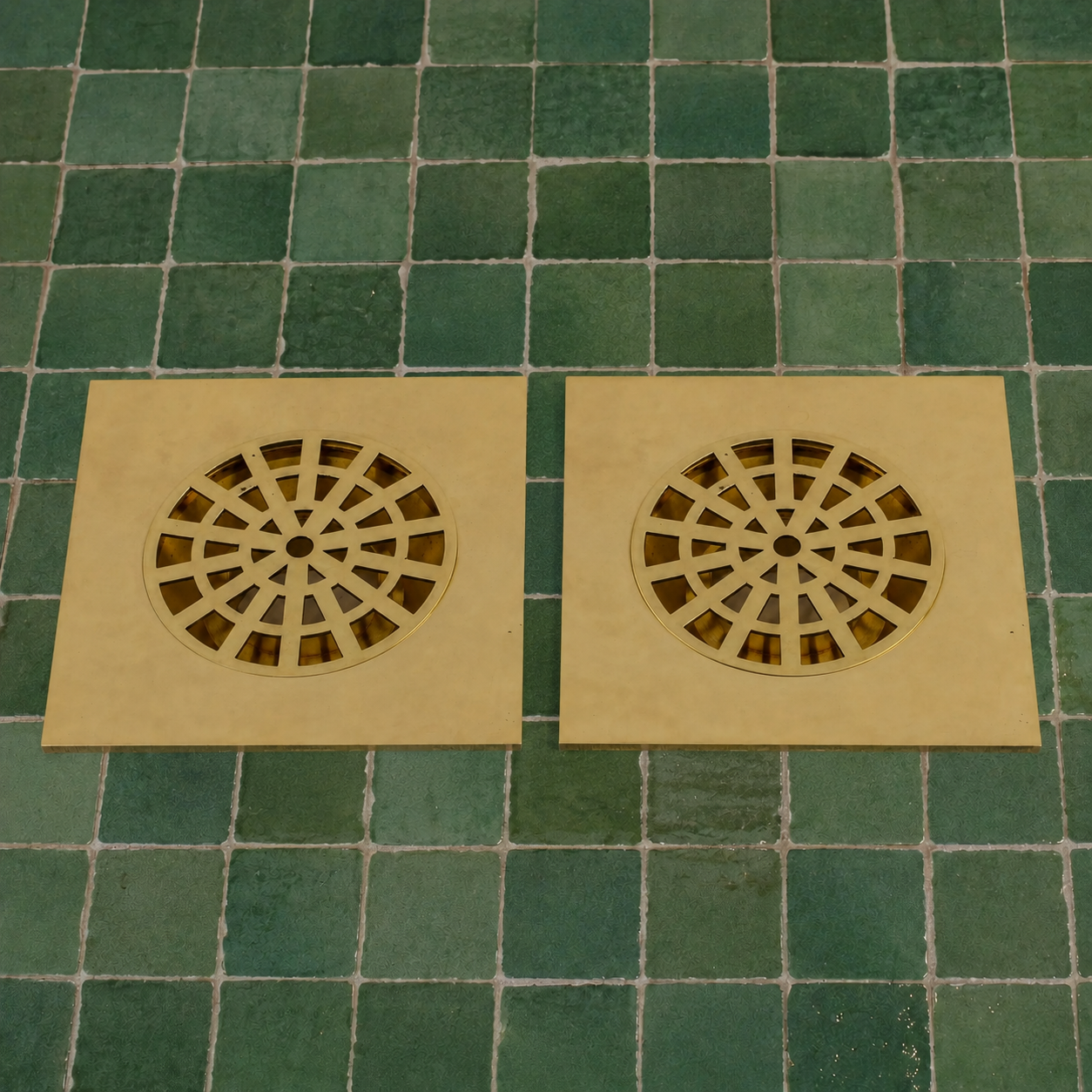 Two square beige drain covers with circular slotted centers, laid side by side on a green tiled floor.