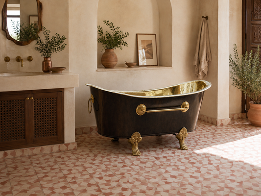 A freestanding clawfoot bathtub with a glossy black exterior and polished gold interior, set in a rustic bathroom with patterned tile flooring, stone walls, and potted plants.
